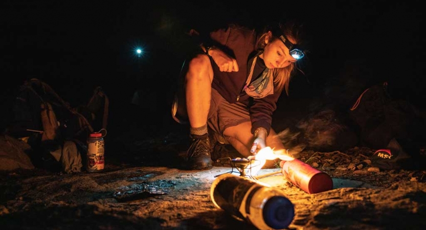 A person wearing a headlamp stokes a small fire while backpacking with outward bound. 